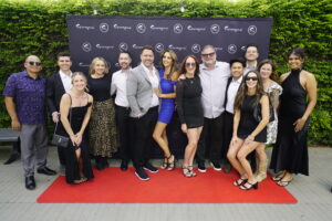 Group of well-dressed people posing on a red carpet in front of a black branding backdrop outdoors, with a green hedge behind them.