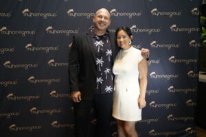 Two people posing together in front of a myhomegroup backdrop, smiling; man in a dark suit with a palm‑tree shirt and woman in a white dress on his side.