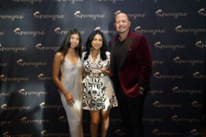 Three people posing for a photo at a My Home Group event, two women in dresses and a man in a burgundy blazer against a branded backdrop